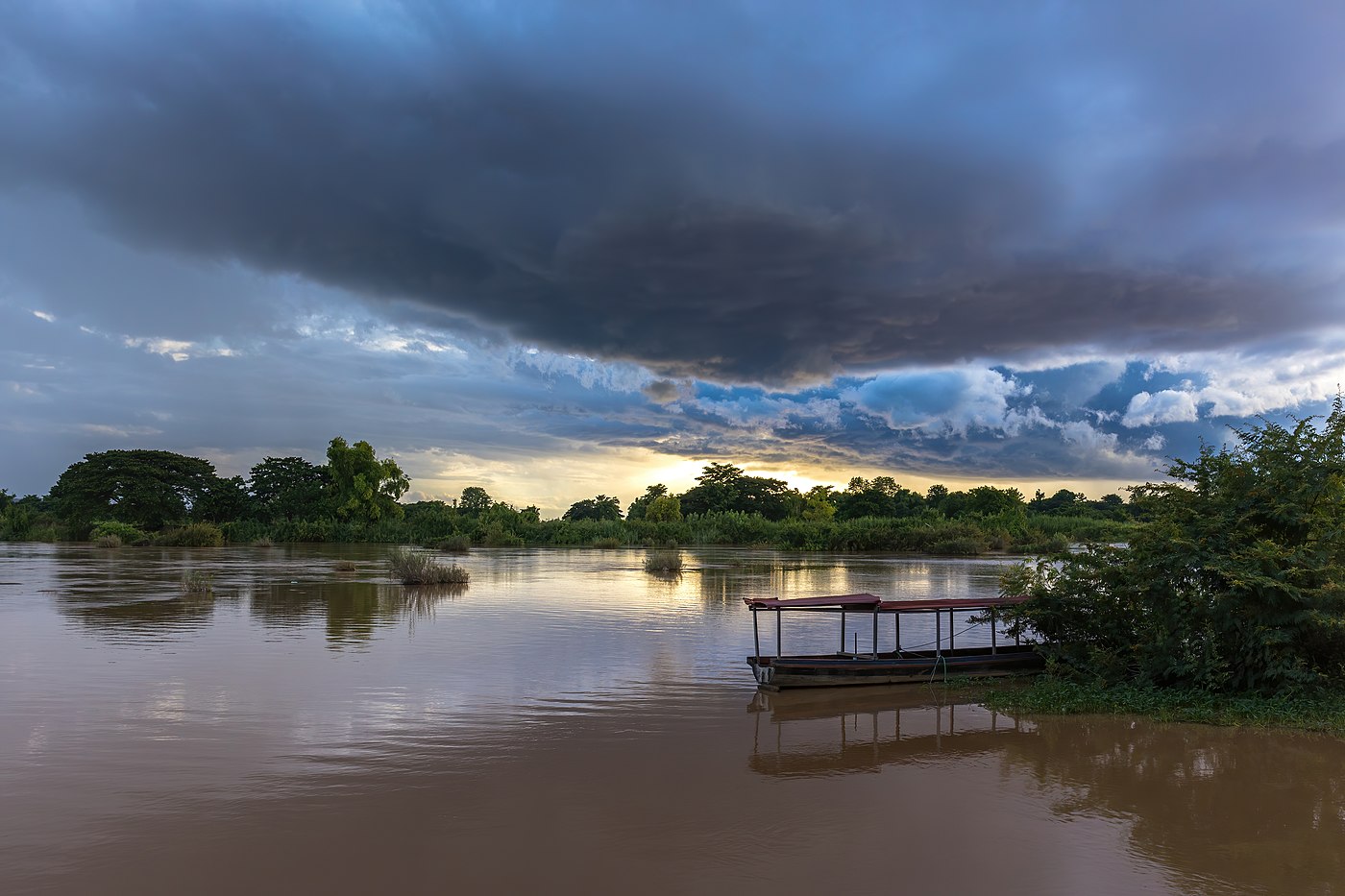 Blue stormy clouds over the Mekong at sunset with water reflection and a pirogue moored to the bank