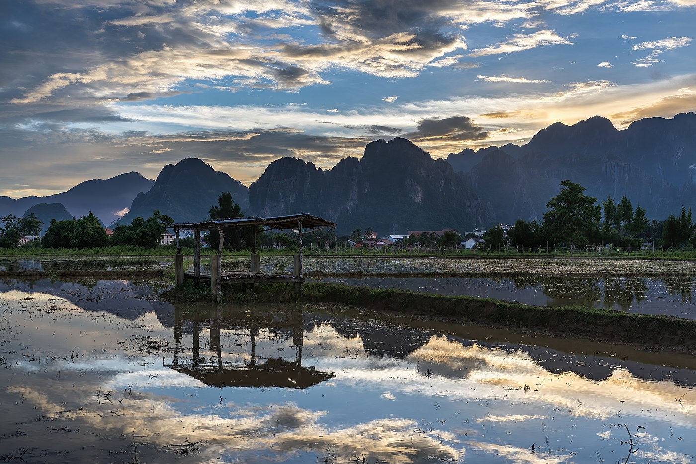 Water reflection of a wooden hut with colorful sky made of orange, gray and white clouds in a paddy field